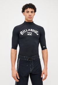 Fekete rövid ujjú rash guard, fehér "BILLABONG" logóval. Fekete fürdőnadrággal párosítva, fehér oldalsávokkal és zsinórral.