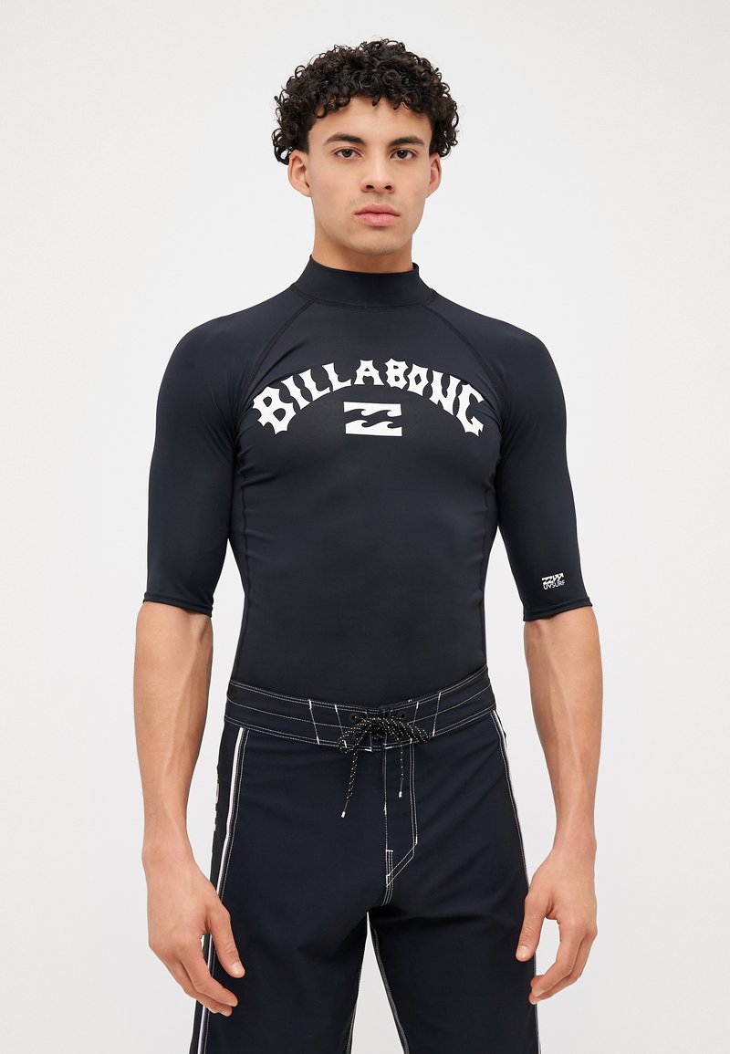 Fekete rövid ujjú rash guard, fehér "BILLABONG" logóval. Fekete fürdőnadrággal párosítva, fehér oldalsávokkal és zsinórral.