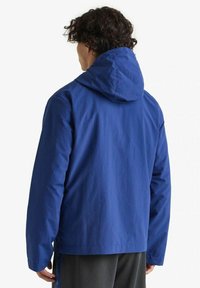 Giacca a cappuccio blu con una texture liscia e leggera. Presenta una vestibilità rilassata, cordoncino in fondo e un design minimalista.