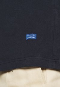 Černé bavlněné tričko s modrou obdélníkovou etiketou s textem "LIBERTINE". Materiál má hladkou strukturu s rovným lemem.