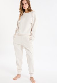 Lichtbeige loungewear-set met een lange mouw top met knoopdetails op de schouder en taps toelopende broek met zijzakken.