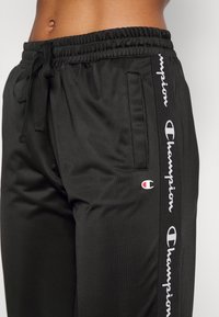 Černé teplákové kalhoty Champion s elastickým pasem, šňůrkou na stažení, bočním zipovým kapsičkou a logem Champion podél pruhu na nohavici.