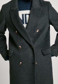 Grijze dubbele rij knoop wollen jas met een geribde kraag, geaccentueerd met bronzen knopen en zijzakken, gelaagd over een blauw gebreide trui met patroon.