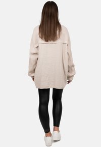Lichtbeige oversized shirt van textiel met structuur, lange mouwen en detail op de rug, gecombineerd met zwarte leggings en witte sneakers.
