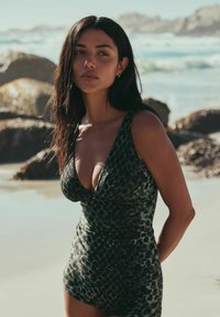 Ung kvinde med langt mørkt hår i en dyb V-hals leopardmønstret badedragt, stående på en solrig strand med klipper og hav i baggrunden.