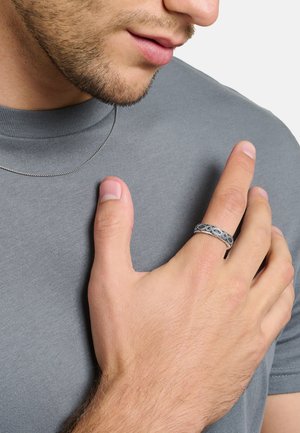 Homme portant une chemise grise et un collier chaîne en argent, touchant sa poitrine avec la main montrant une bague en argent détaillée sur le majeur.