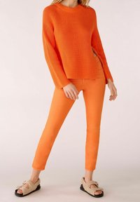 Femme debout portant un pull en tricot orange et un pantalon orange, chaussant des sandales ouvertes beiges en peluche avec des semelles noires, sur un fond uni.