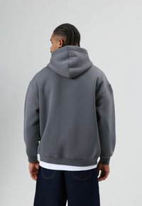 Grijze capuchontrui met een ontspannen pasvorm en geribde manchetten. Heeft een zachte textuur en een buidelzak, gedragen over donkere spijkerbroek.