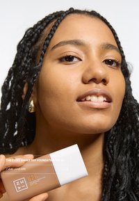 Fundamentfles met het label "Tan warm, 626" met een glanzend beige body en een witte dop, vastgehouden door een persoon met vlechten en een egale huid.