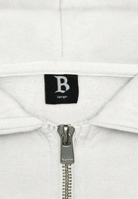 Haut de sweatshirt à zip blanc avec une doublure intérieure douce, doté d'une fermeture éclair argentée et d'une étiquette noire portant la mention "B large". Tissu texturé avec un design simple.