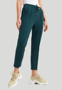 Tyrkysové tepláky s páskem na stahování, slim fit a prošitím, spárované s světle béžovými sportovními teniskami.