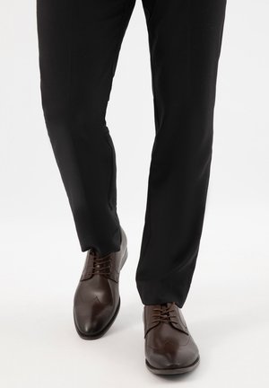 Donkere bruine leren herenschoenen met brogue-details, gedragen met zwarte broek, op witte achtergrond.