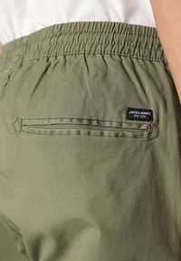 Olijfgroene broek met een elastische tailleband, voorzien van een achterzak en een zwart label met witte tekst, gemaakt van gestructureerde stof.