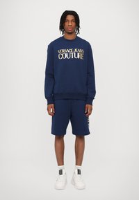 Temno modra majica s kapuco z napisom "VERSACE JEANS COUTURE" v zlatih črkah, v kombinaciji z ujemajočimi kratkimi hlačami. Ima ribane manšete in stranske dodatke.