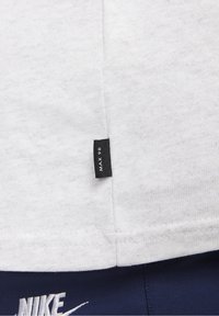 Vit bomull t-shirt med en texturerad yta, med en liten svart etikett som visar "MAX 90" på sidsömmen. Marinblått tyg synligt nedanför.