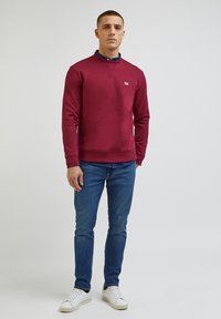 Sweatshirt bordeaux com gola redonda, punhos e bainha canelados. Combinado com jeans azuis e ténis brancos. Pequeno detalhe de logótipo no peito.