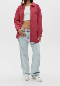 Veste rouge boutonnée avec une poche poitrine unique, portée sur un débardeur blanc, associée à un jean large bleu clair et des baskets blanches.