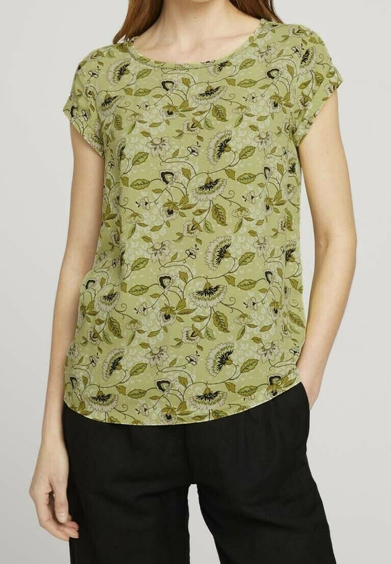 Femme portant un chemisier à manches courtes floral vert clair associé à un pantalon noir, debout avec une main dans la poche.