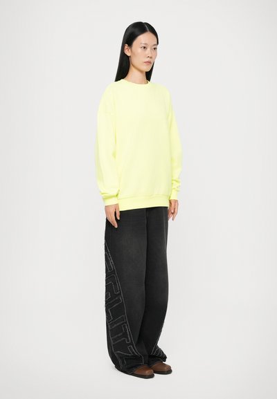 Neon gul oversized sweatshirt med løstsiddende sorte denimbukser med et tekstureret, iøjnefaldende logodesign på siden. Brune slip-on sko.