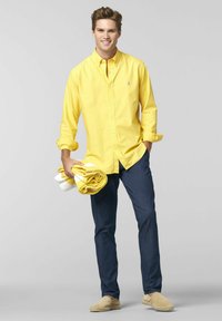 Gul button-up skjorte med lange ærmer, mørkeblå jeans og lysebeige espadrillos. Holder rullede håndklæder i gul og hvid.