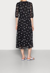 Schwarzes Midi-Kleid mit weißen Blumenmustern, durchsichtigen Puffärmeln, gerafftem Taillenbereich und fließendem Rock. Kombiniert mit helllila Sling-Sandalen.
