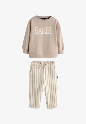 Giovane ragazzo sorridente che indossa una felpa beige "BAKER", pantaloni a righe e sneakers bianche, in piedi con le mani parzialmente nelle tasche.