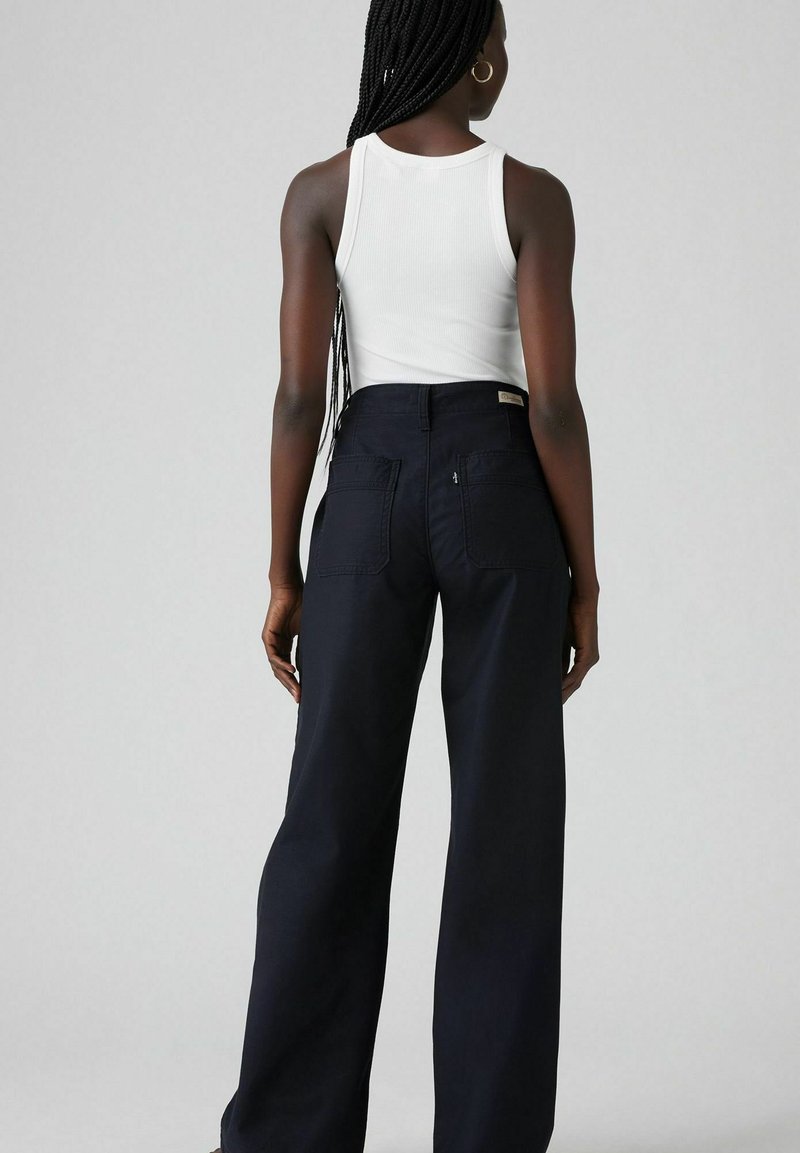 Pantaloni neri a gamba larga con due tasche posteriori, abbinati a un top bianco senza maniche. Realizzati in un tessuto liscio e resistente.