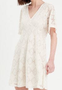 Robe en dentelle couleur crème avec un décolleté en V et des manches courtes, présentant des motifs géométriques complexes et une silhouette évasée.