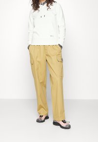 Felpa bianca con cappuccio e coulisse, con tasca frontale, abbinata a pantaloni cargo color beige con tasche laterali e etichetta nera, indossata con scarpe sportive.