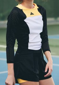 Svart och gul atletisk skjorta med långa, perforerade ärmar och en vit frontpanel, tillsammans med en svart kjol. Texturerat material, åtsittande design.