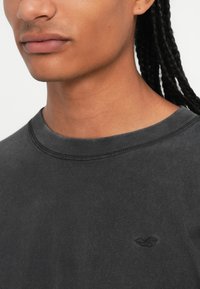 Tamno siva pamučna majica s okruglim izrezom i kratkim rukavima, s diskretnim crnim logotipom izvezenim na prsima.