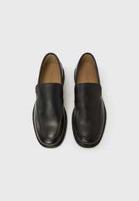 Zapatos negros de cuero sin cordones con textura suave, punta redondeada y costuras decorativas. Plantilla acolchada visible en el interior.