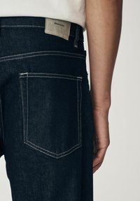 Sötét farmernadrág egyenes szabással, kontrasztos fehér varrással. Egy kis bézs címke látható a deréknál, amelyen az áll: "DENIM SUPPLY".
