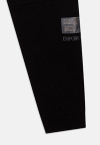 Des joggers en coton noirs présentent un patch en argent texturé avec un motif géométrique et un logo brodé de la marque sur la jambe inférieure.