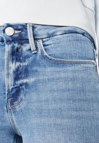 Gros plan sur la taille d'un jean en denim bleu clair avec bouton métallique, passants pour ceinture, poche avant et partie d'une chemise blanche rentrée.