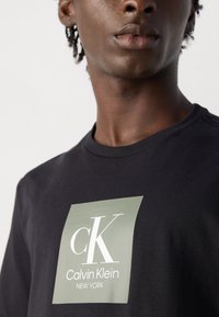 Acercamiento a una persona que lleva una camiseta negra de Calvin Klein con un logo cuadrado gris y el texto blanco "CK Calvin Klein New York".