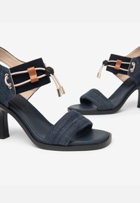 Denim højhælede sandaler med en sort sål, frontrem og ankelstrop med tan detalje og snoredekurv. Struktureret mørkeblå overflade.