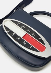 Granatowa torebka syntetyczna z dużym srebrnym owalnym logo z czarnymi, białymi i czerwonymi sekcjami, wytłoczonym napisem "TOMMY JEANS."