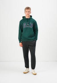 Green hoodie with "GAP" printed in large letters, paired with gray sweatpants. Model wears beige sneakers. Simple design, casual fit.