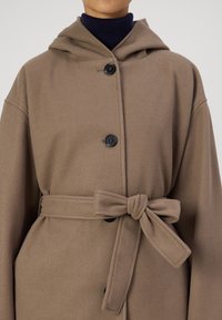 Manteau en laine marron avec une capuche, doté de deux boutons noirs et d'une ceinture nouée à la taille. Texture douce et coupe ample.