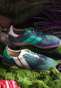 Paire de baskets en daim bleu sarcelle et vert avec des bandes contrastantes, posées sur de la mousse et entourées de plantes dans un cadre forestier.