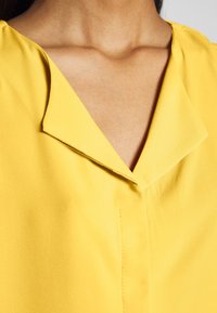 Donna che indossa una blusa gialla con un ampio colletto a V pieghe, che mostra parte del suo collo e della parte superiore del petto.