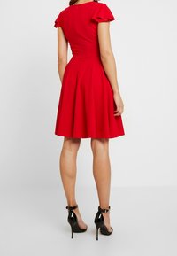 Czerwona sukienka z krótkimi, falbaniastymi rękawami, dopasowanym gorsetem i rozkloszowaną spódnicą. Noszona z czarnymi, paskowymi sandałami na wysokim obcasie. Gładka faktura.