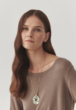 Gold chain necklace with a green and white marbled pendant, worn with a light brown sweater. Hair is long and wavy. Neutral background.