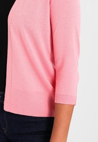 Jasnoróżowy kardigan z rękawami do łokcia, ściągaczami na mankietach i dolnym brzegu, ozdobiony subtelnym perforowanym wzorem wzdłuż szwów.