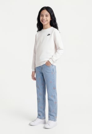 Usmievavá dievčina s dlhými čiernymi vlasmi, oblečená v bielej mikine Nike, svetlomodrých džínsoch s malými oranžovými vzormi a bielych teniskách, stojaca.