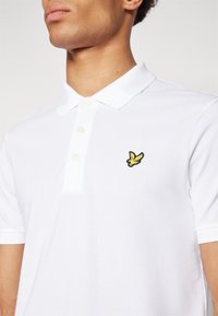 Polo blanc en tissu texturé, doté d'une patte à deux boutons et d'un petit logo brodé d'un oiseau jaune sur la poitrine gauche.
