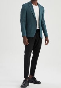 Blazer di misto lana color teal con revers a punta, due tasche frontali, chiusura a un bottone, abbinato a pantaloni neri a gamba affusolata e scarpe nere.