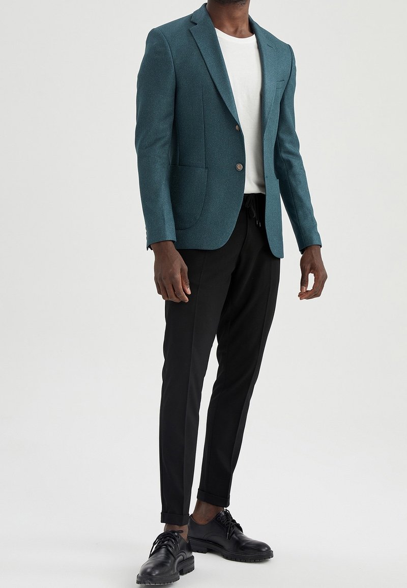 Blazer di misto lana color teal con revers a punta, due tasche frontali, chiusura a un bottone, abbinato a pantaloni neri a gamba affusolata e scarpe nere.