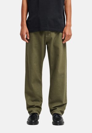 Personne portant un pantalon ample vert olive, des chaussures noires et une chemise noire, debout avec les mains détendues le long du corps.